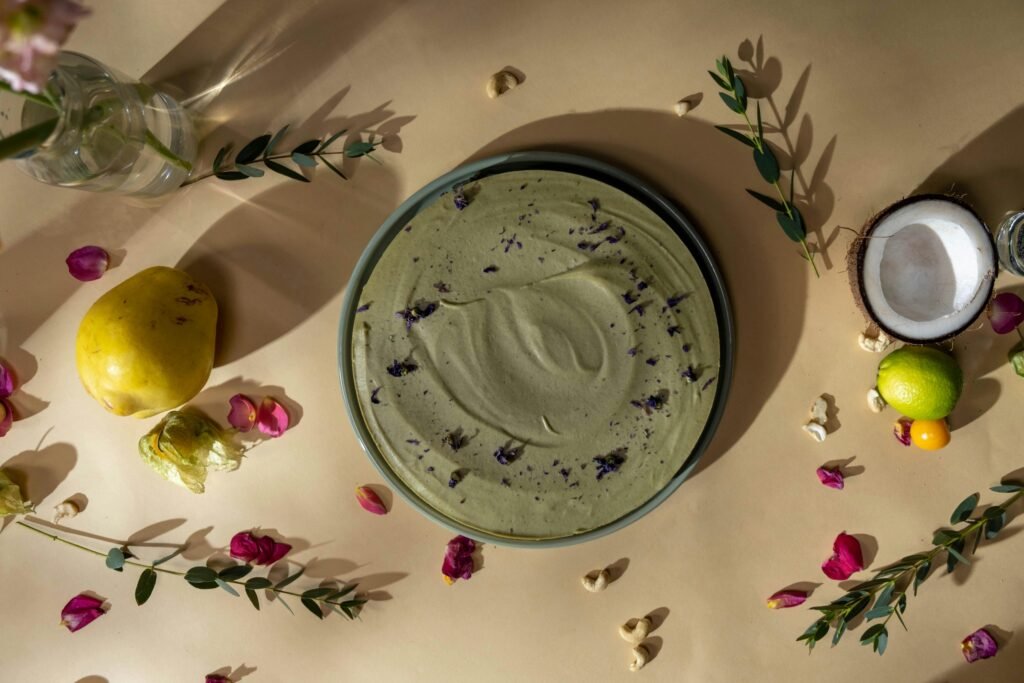 Top-down view of a vegan green cake garnished with flower petals, surrounded by fruits and leaves.